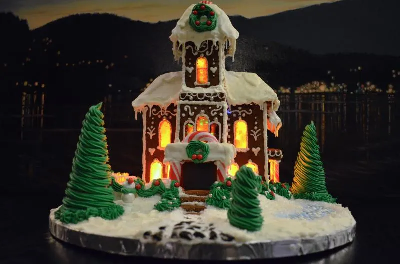 Lit gingerbread cottage with a wreath on its peak and green frosted trees on a round snowy base
